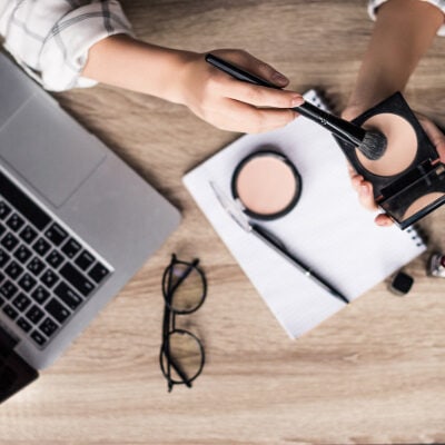 overhead shot of desk; woman applies makeup with brush while laptop, notebook, and glasses are also on desk
