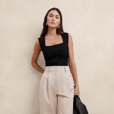 A woman wearing a black sleeveless top, khakis, and gold earrings, and carrying a black handbag
