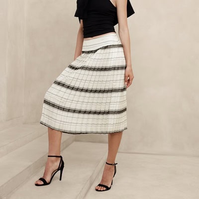 A woman wearing a black sleeveless top, another black top over her shoulders, a black-and-white striped skirt, and black sandals