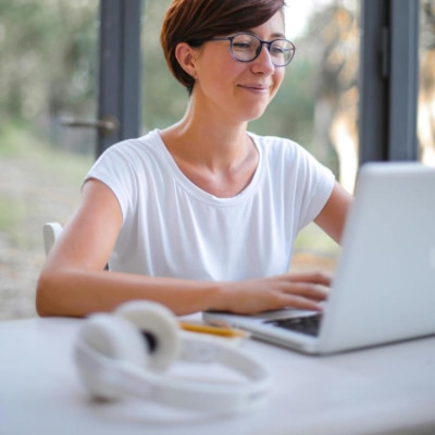 woman wears an opaque white t-shirt for women; she is typing on a laptop and sitting in front of a window with trees and grass behind her; in front of her on the desk sits a pair of white head phones.