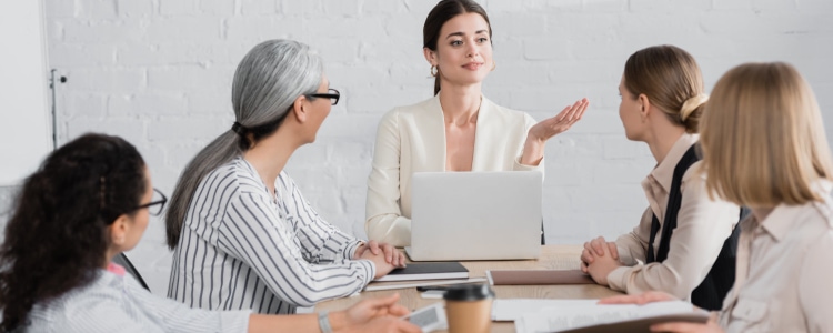 young professional woman sits at head of table and gestures with her hand; she has a laptop in front of her. The rest of the table has other women executives.