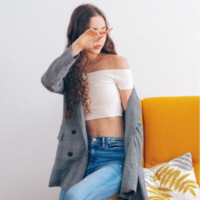 woman wears a white crop top, blue jeans, and a blazer; she sits on the edge of a yellow couch