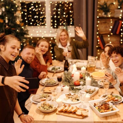 A young woman stands at a dinner table, taking a selfie or movie of the group; the other dinner guests are waving and smiling in the background.