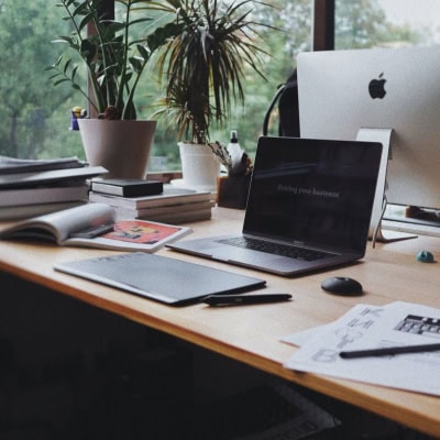 laptop sits on desk in front of window, surrounded by books, plants, and an open magazine