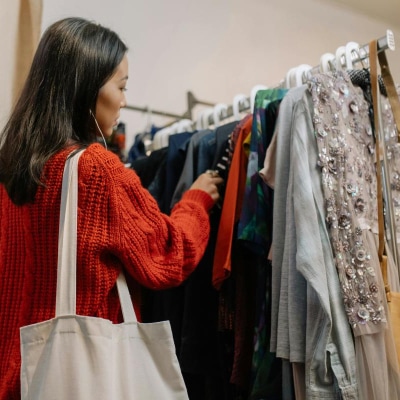 woman looks through a thrift store rack; she is trying to make her workwear more eco-friendly. She is wearing a red sweater and has a cream canvas bag slung over her shoulder.