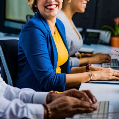 smiling woman sits at conference table with other people; she wears red lipstick, a yellow t-shirt and a cobalt blue blazer