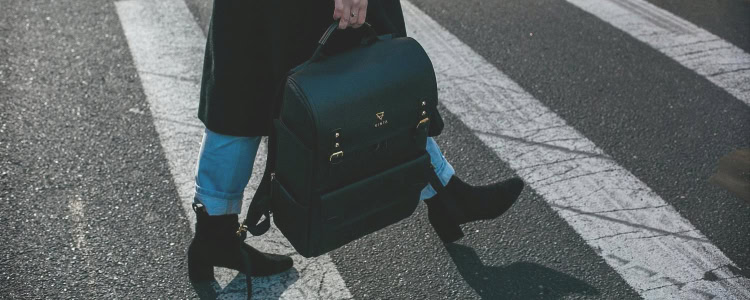 young woman walking to her casual office; she is wearing blue jeans, black boots, a black overcoat, and carrying a large structured tote/briefcase bag. 
