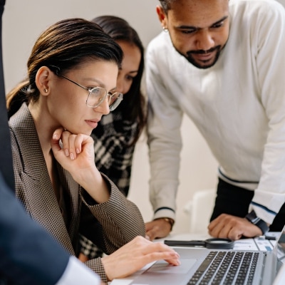 young professional woman wears makeup with eyeglasses; she is looking at a laptop screen with two colleagues next to her, one with a beard and mustache and another with longer hair