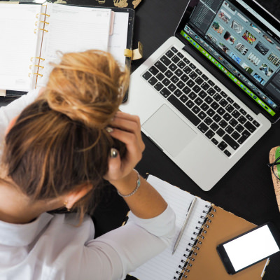 a woman with her head in her hands; she has a messy bun and is sitting in front of a laptop and a notebook