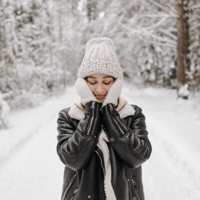 woman pats her face; she is wearing beige gloves and a hat (and a black leather jacket); in the background you can see trees covered in snow