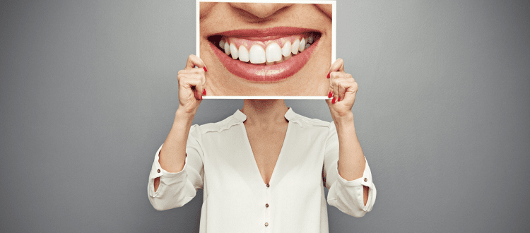 Professional woman wearing a white blouse, holding up a large photograph of a woman's mouth smiling -- the photograph is so large it covers the woman's head entirely