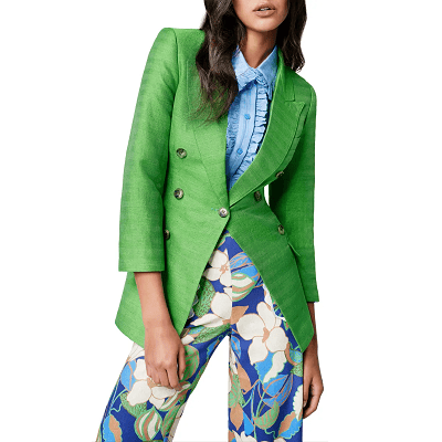 woman slouches in one of the best summer blazers, a green linen-silk option from Smythe -- she is also wearing colorful floral pants and a ruffly blue blouse