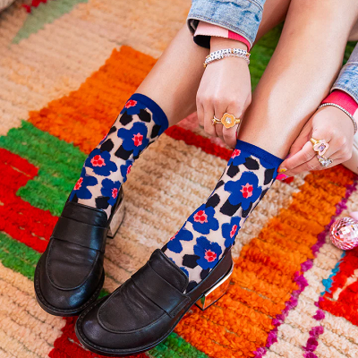 woman wears sheer socks with colorful pattern in blue, black, pink, and red; she is sitting on a colorful rug with oranges, greens, and reds